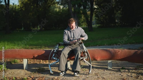 Young man in gray hoodie sitting on park bench adjusting metallic spring stilts on sunny day, surrounded by green grass and trees, with focused expression in peaceful outdoor setting photo