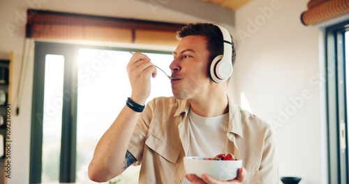 Headphones, eating and man with breakfast in kitchen listening to podcast in morning for self care. Audio tech, health and male person enjoying fruit salad for diet, nutrition or wellness in home. photo