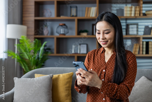 Asian woman smiling and using smartphone at home. Seated on sofa in modern living room, engaged in social media browsing or chatting. Captures moments of relaxation and connectivity.