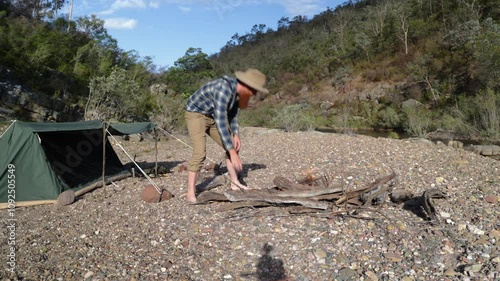 Bushman in Australia lights a campfire by a river on a sandy bank with a canvas tent. photo