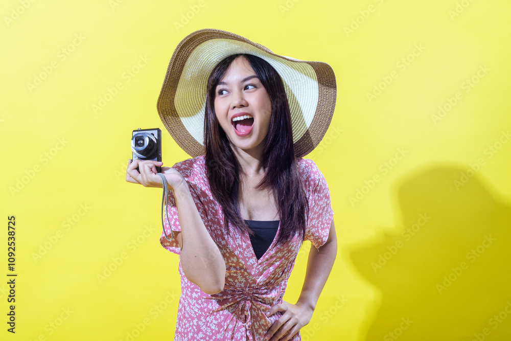 Indonesian Asian woman in a straw hat and pink floral dress, holding a camera and looking to the side with an excited, open-mouthed smile, against a yellow background. Bright, even lighting Indonesian Asian woman in a straw hat and pink floral dress, holding a camera and looking to the side with an excited, open-mouthed smile, against a yellow background. Bright, even lighting