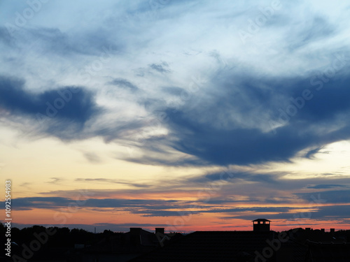 The evening sky glows with a mesmerizing blend of orange, blue and purple hues as the sun sets. Silhouettes of houses stand peacefully beneath, their roofs bathed in the fading sunlight.