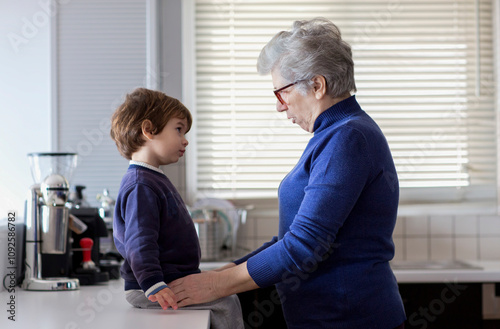 GraA grandmother and her young grandson share a moment in a bright, modern kitchen, engaging in conversation. The child looks up at her attentively while she kneels beside him, fostering connection. photo