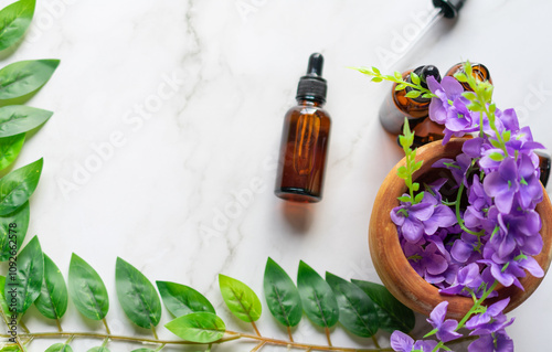 A mortar with some purple flowers and some amber vials of essential oils on a white marble surface. Leaves covering the opposite corners photo