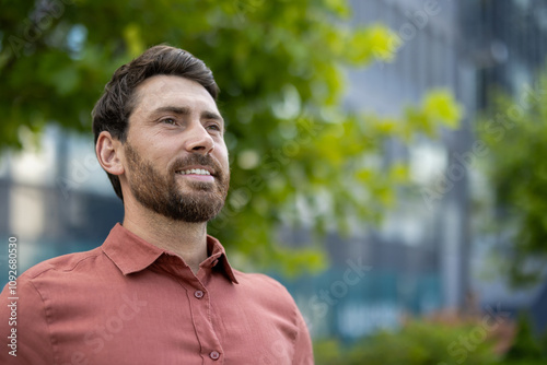 Mature businessman wearing casual shirt smiling outdoors with green leaves and office buildings background. Represents confidence, success, and career growth in a professional setting.