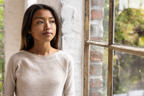 Asian female teenager standing by window, looking outside thoughtfully at home photo