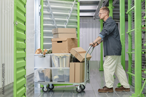 Full length shot of handsome man loading cart with cardboard boxes into self storage unit at warehouse while moving out or decluttering apartment photo