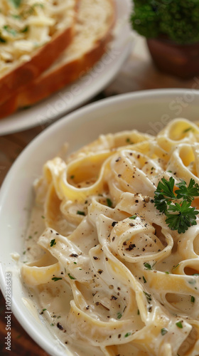 Vertical close-up of Fettuccine Alfredo with creamy sauce enveloping the pasta, garnished with parsley and black pepper, and a slice of garlic bread in the background. photo