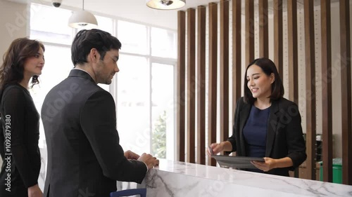 Businessman and woman with luggage check in at the hotel reception lobby, female receptionist welcoming guest and counter desk