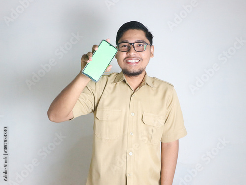 An Asian male Indonesian civil servant wearing a brown uniform, usually called a keki, stands while holding a cellphone while showing a blank or greenscreen. Indonesian civil servants, ASN photo