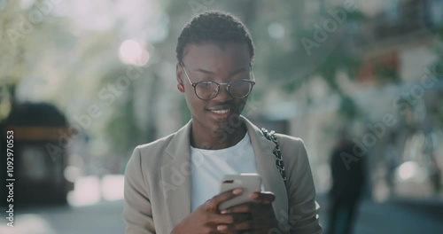 Smiling young African American woman with glasses walking use phone in city center. Businesswoman telephone. Messaging outdoor smartphone. Girl, texting. Slow motion