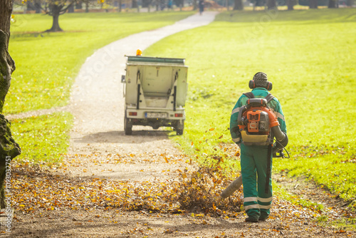 Back view of a public worker using leaf blower to clean path in a park from fallen leaves. Autumn scene in park, maintenance of paths, utility vehicle visible...