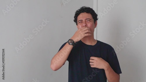 Man Sneezing into Elbow. A man in a black t-shirt standing in front of a white wall. He is looking at the camera.