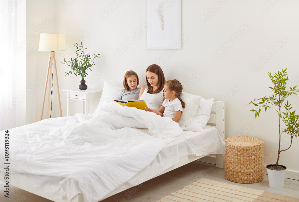 Happy family mother and two kids lying in comfortable bed under blanket, reading interesting book together. Caring mom reading fairytale to her children, enjoying leisure time during weekend together. Happy family mother and two kids lying in comfortable bed under blanket, reading interesting book together. Caring mom reading fairytale to her children, enjoying leisure time during weekend together.