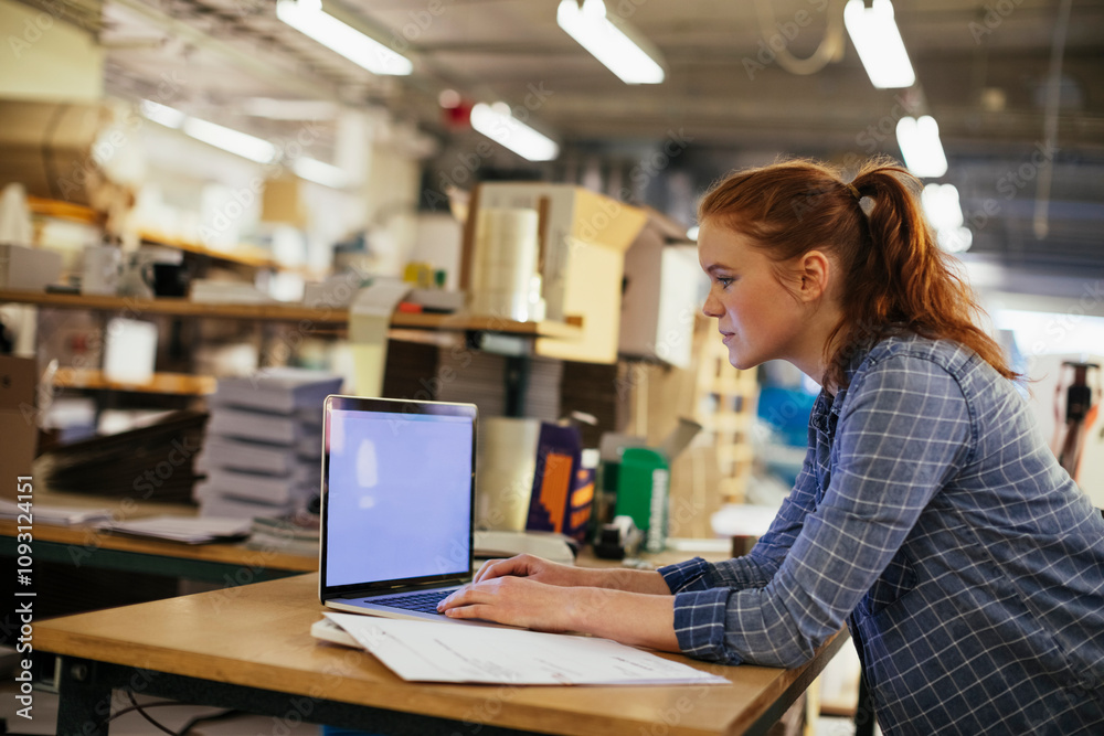 Young woman using a laptop while working in a printing press office Young woman using a laptop while working in a printing press office