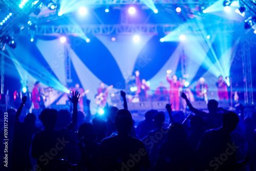 Silhouettes of crowd at concert in front of stage with bright spotlights.The back view of people enjoy live music at the band's performance. Outdoor concert with musicians on stage and fans.