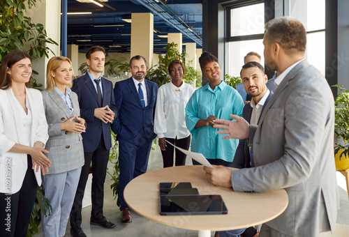 Diverse group of company employees stand in row, listening intently to experienced male business leader or coach during staff meeting or workshop. Trainer talking about project, sharing his expertise.