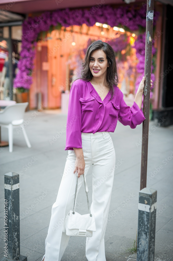 Young beautiful brunette woman in a pink jacket and white pants posing on the street. Portrait of a beautiful young woman in a purple blouse and white pants posing in an outdoor cafe on the street