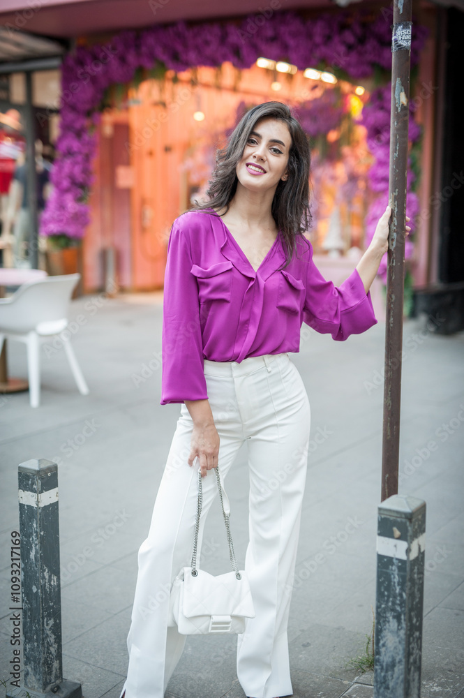 Young beautiful brunette woman in a pink jacket and white pants posing on the street. Portrait of a beautiful young woman in a purple blouse and white pants posing in an outdoor cafe on the street