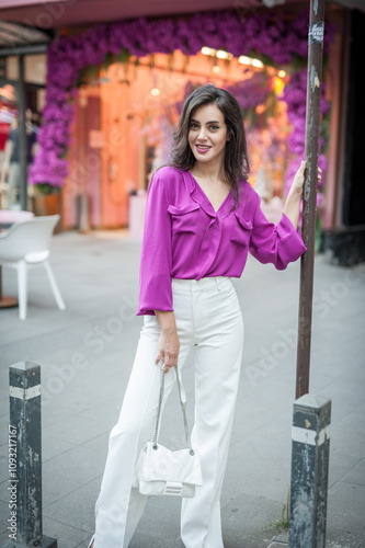 Obraz na płótnie Young beautiful brunette woman in a pink jacket and white pants posing on the street. Portrait of a beautiful young woman in a purple blouse and white pants posing in an outdoor cafe on the street w salonie