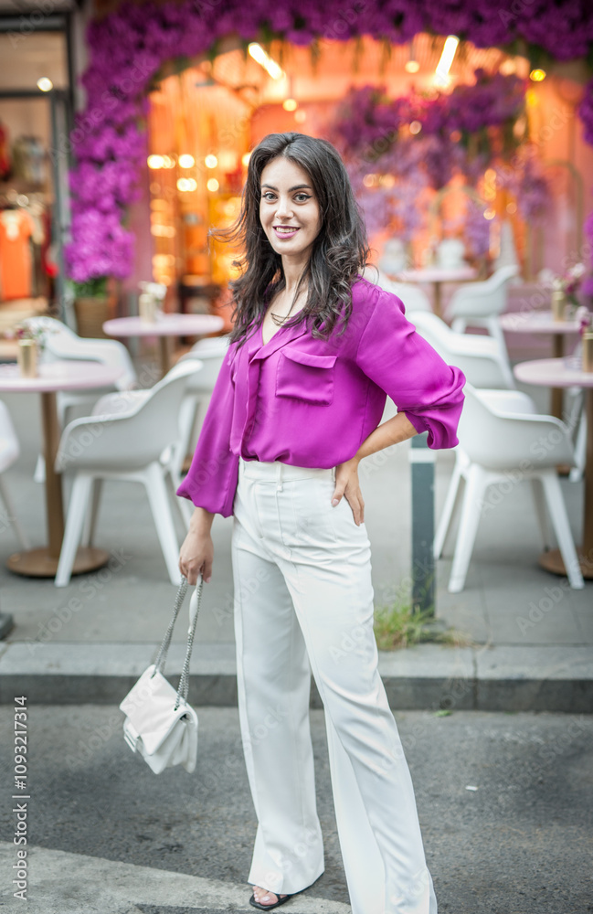 Young beautiful brunette woman in a pink jacket and white pants posing on the street. Portrait of a beautiful young woman in a purple blouse and white pants posing in an outdoor cafe on the street