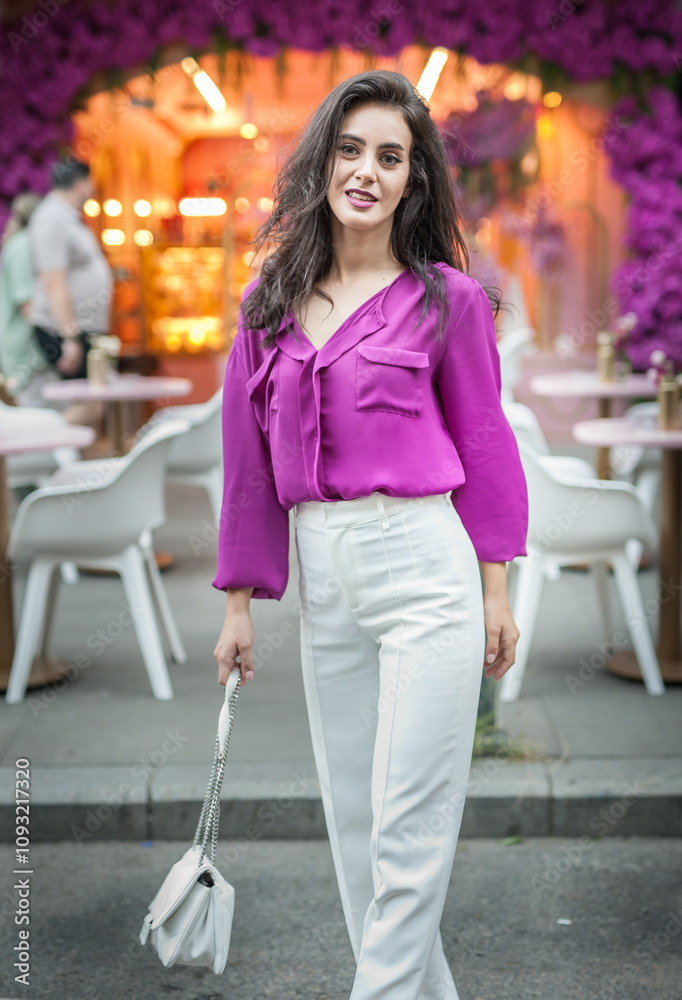 Young beautiful brunette woman in a pink jacket and white pants posing on the street. Portrait of a beautiful young woman in a purple blouse and white pants posing in an outdoor cafe on the street