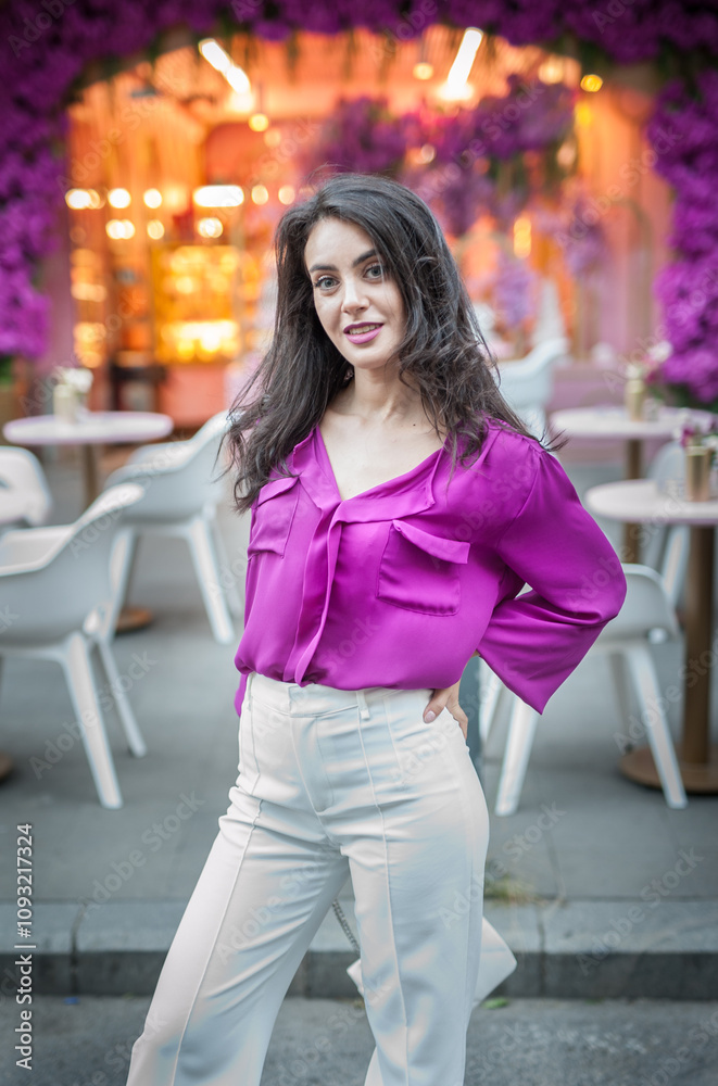 Young beautiful brunette woman in a pink jacket and white pants posing on the street. Portrait of a beautiful young woman in a purple blouse and white pants posing in an outdoor cafe on the street