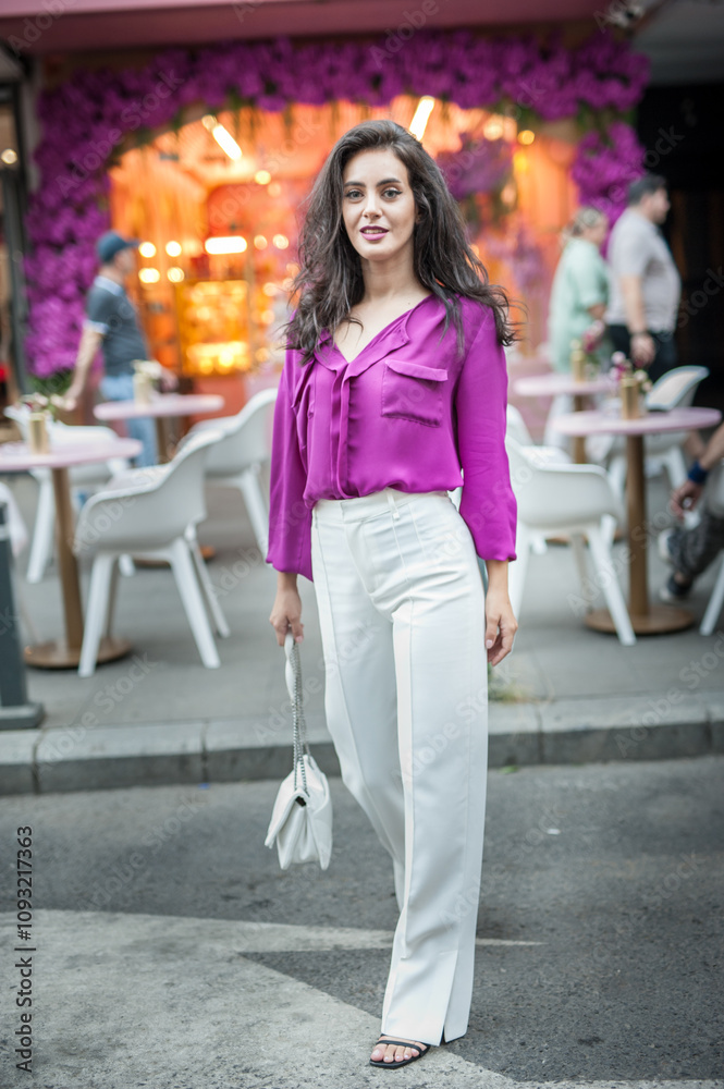 Young beautiful brunette woman in a pink jacket and white pants posing on the street. Portrait of a beautiful young woman in a purple blouse and white pants posing in an outdoor cafe on the street