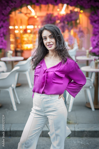 Obraz na płótnie Young beautiful brunette woman in a pink jacket and white pants posing on the street. Portrait of a beautiful young woman in a purple blouse and white pants posing in an outdoor cafe on the street w salonie