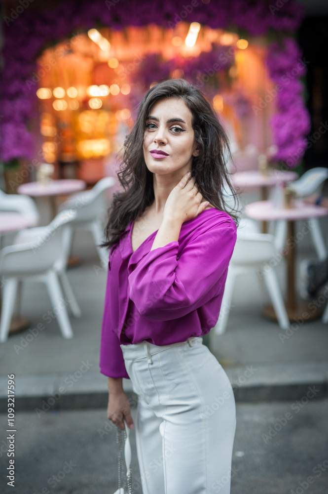 Young beautiful brunette woman in a pink jacket and white pants posing on the street. Portrait of a beautiful young woman in a purple blouse and white pants posing in an outdoor cafe on the street
