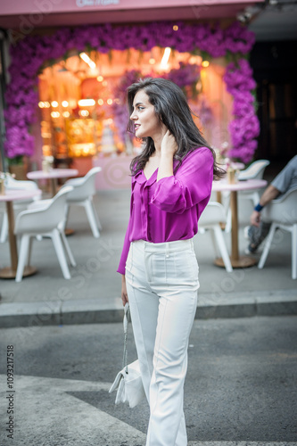 Young beautiful brunette woman in a pink jacket and white pants posing on the street. Portrait of a beautiful young woman in a purple blouse and white pants posing in an outdoor cafe on the street