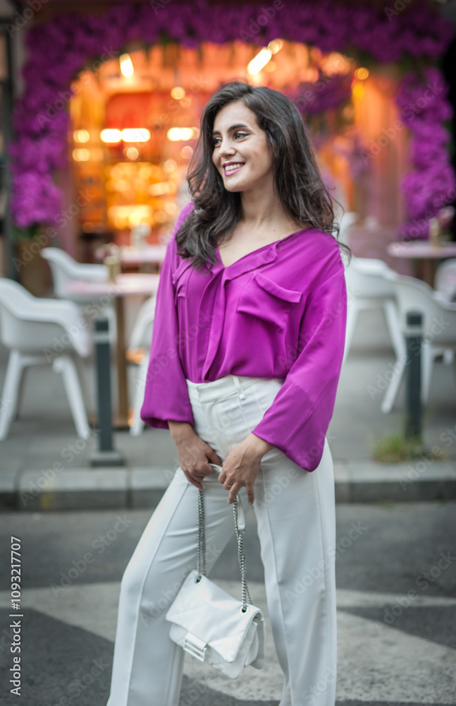 Young beautiful brunette woman in a pink jacket and white pants posing on the street. Portrait of a beautiful young woman in a purple blouse and white pants posing in an outdoor cafe on the street