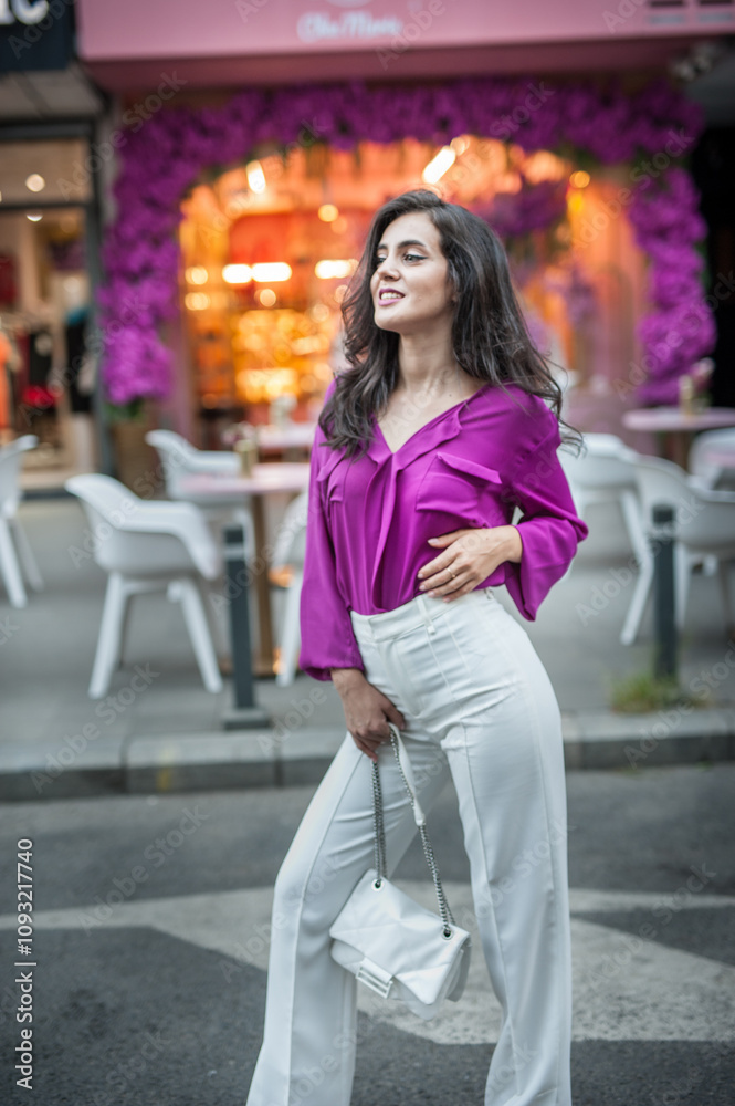 Young beautiful brunette woman in a pink jacket and white pants posing on the street. Portrait of a beautiful young woman in a purple blouse and white pants posing in an outdoor cafe on the street