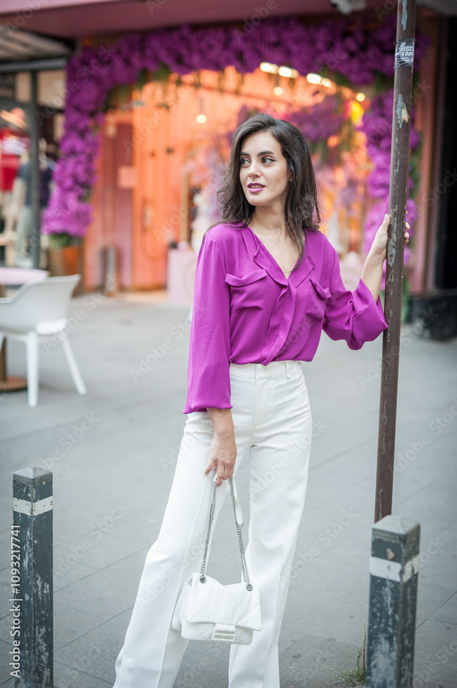 Young beautiful brunette woman in a pink jacket and white pants posing on the street. Portrait of a beautiful young woman in a purple blouse and white pants posing in an outdoor cafe on the street