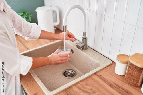 Woman fills a glass with clean filtered tap water for drinking in scandinavian style kitchen at home