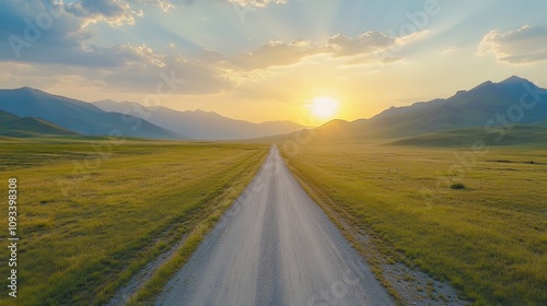 Serene sunrise view of an empty road flanked by lush grassy fields and majestic mountains under a colorful sky, evoking tranquility in nature.