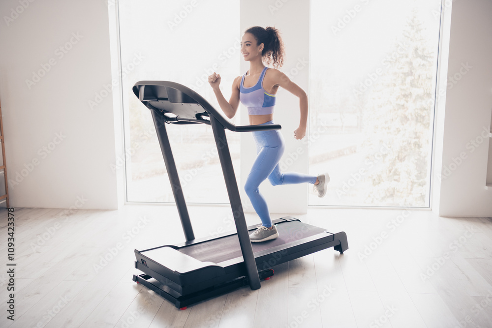Young woman enjoys fitness session on treadmill in sunny modern home interior, wearing trendy sportswear