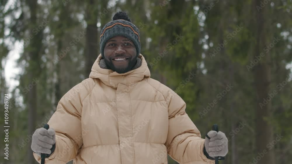 Waist up portrait of optimistic Black young man dressed in winter clothes posing for camera with ski poles in woods Waist up portrait of optimistic Black young man dressed in winter clothes posing for camera with ski poles in woods