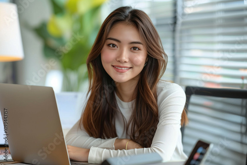 Young Asian Woman Smiling Warmly at Her Workspace with a Laptop in a Modern Office Setting