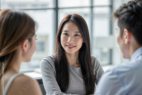 Free Download young Asian woman seated in a bright, modern office smiling at two people, a man and a woman, who are seated across from her. - GFXLoad