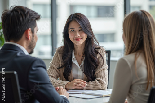 young Asian woman seated in a bright, modern office smiling at two people, a man and a woman, who are seated across from her.