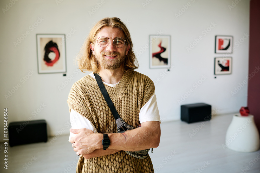 © AnnaStills - Portrait of man with long hair standing in modern art gallery, smiling and wearing casual attire arm crossed and looking directly at the camera