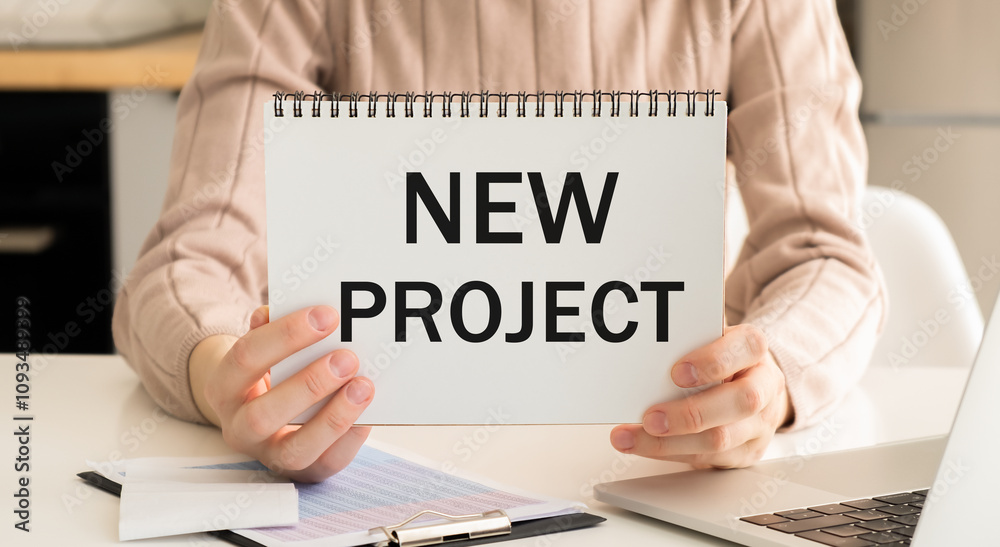 A notebook on a spring with the text NEW PROJECT on a white sheet lies on a brown wooden table with colored pens. A notebook on a spring with the text NEW PROJECT on a white sheet lies on a brown wooden table with colored pens.