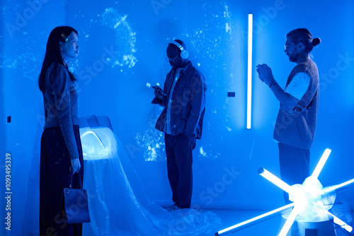 Participants engaging with futuristic techno art installation illuminated by blue lighting. Diverse group examining interactive displays with immersive surroundings