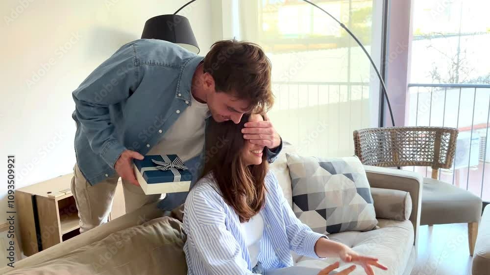 A loving boyfriend surprises his girlfriend sitting comfortably on a sofa by softly covering her eyes and handing her a beautifully wrapped gift, evoking a heartfelt and joyful reaction