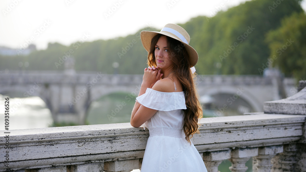 Fototapeta premium An elegant tourist wearing a white dress and straw hat admires the beauty of the Tiber River and a historic Roman bridge.