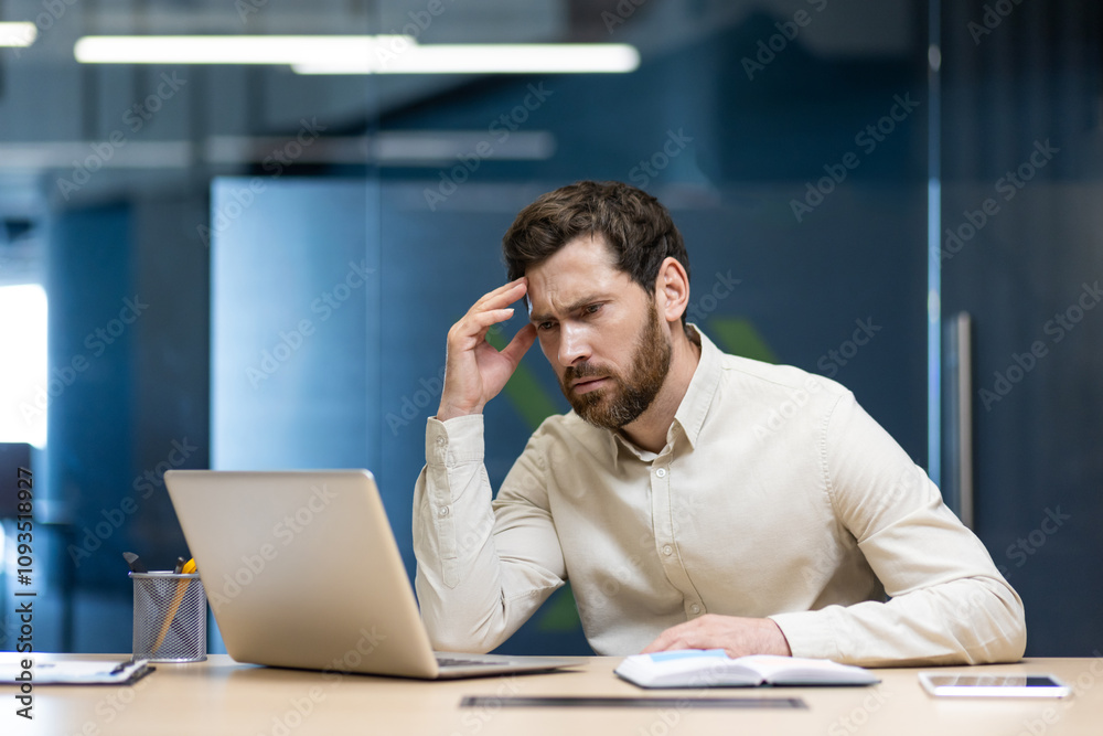 Confident young man sitting in the office at the desk, holding his head and looking worriedly at the laptop Confident young man sitting in the office at the desk, holding his head and looking worriedly at the laptop