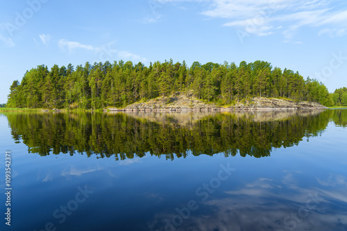 Fototapeta Naklejka Na Ścianę i Meble -  A stone island covered with pine forest is reflected in the smooth water of the lake on a sunny day. Landscape.