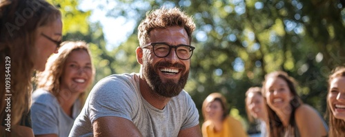 Bearded man with glasses laughing surrounded by a group of friends in a park on a sunny day.
