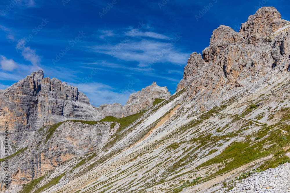 Fototapeta premium Sunny day in the Dolomites. Rocky mountain peaks of Dolomiti Alps in summer under the blue sky with some clouds prominent mountain summits landscape and stunning views of the alpine range and meadow
